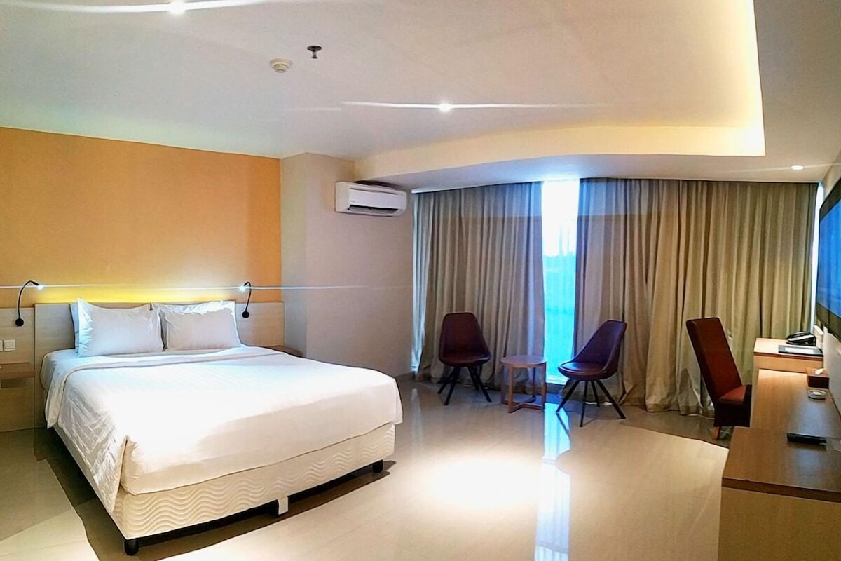 Une chambre d’hôtel avec un grand lit, deux chaises, un bureau et une fenêtre avec des rideaux.