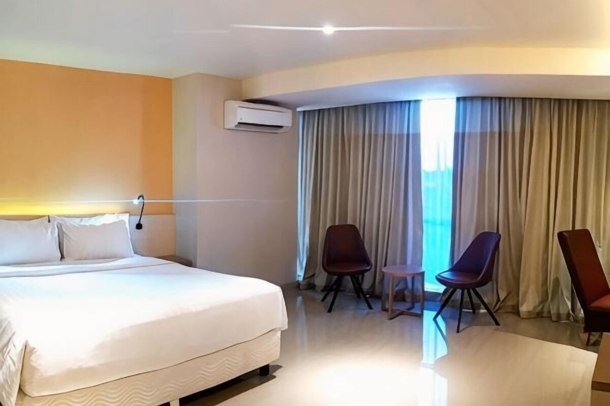 Une chambre d’hôtel avec un lit, deux chaises, une petite table et une grande fenêtre avec des rideaux.