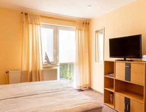 Ein Hotelzimmer mit Bett, Fernseher auf einem Holzständer, einem Fenster mit Vorhängen und Blick ins Grüne.