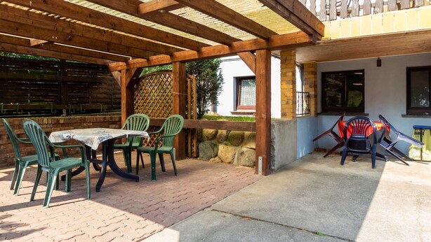 Ein überdachter Patio-Bereich mit Tisch und Stühlen, eine Holz-Pergola und Blick auf ein Gebäude mit Fenster und Tür.