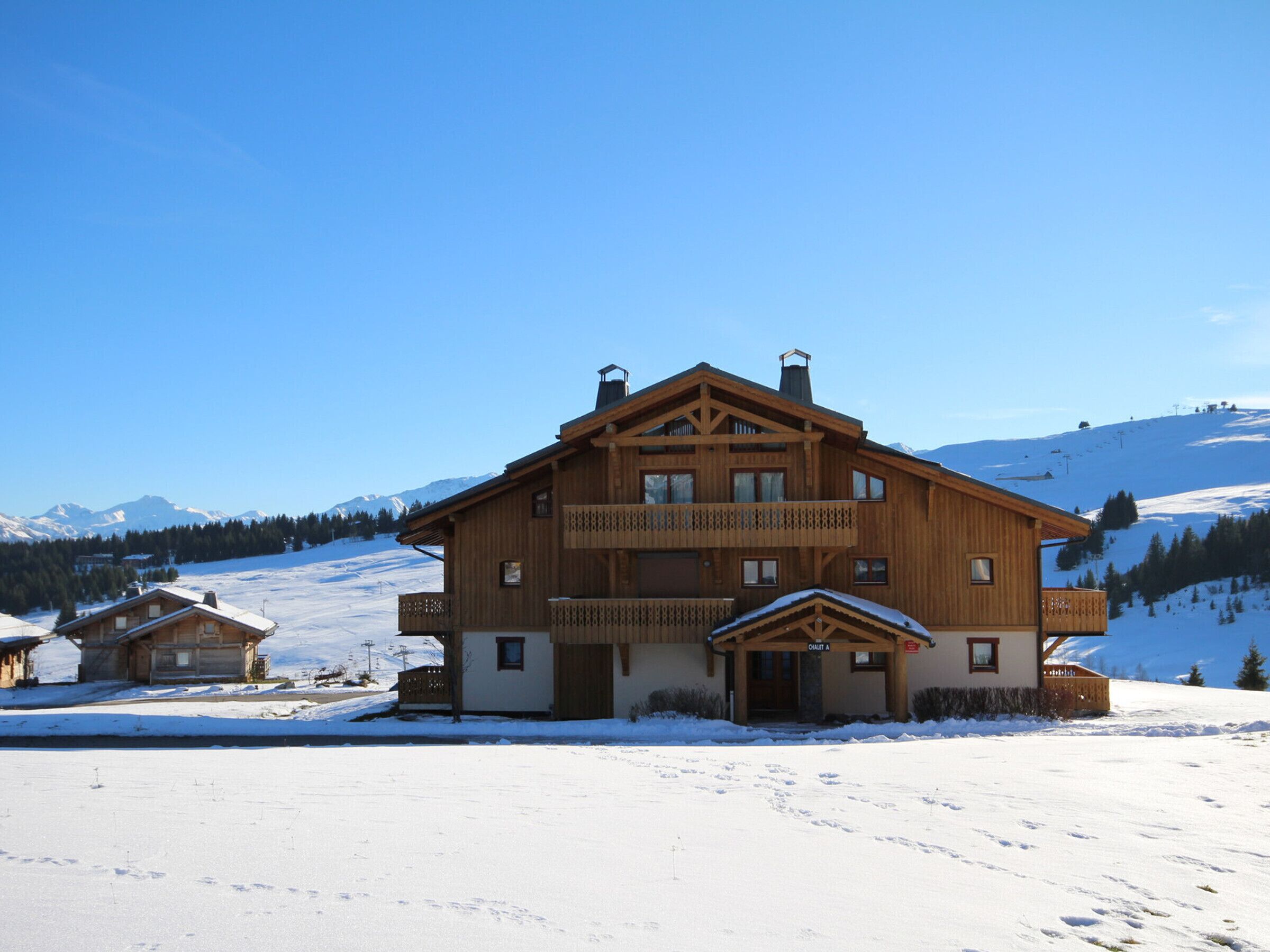 A large wooden chalet with a balcony, situated on a snow-covered slope with mountains in the background.