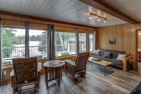 A cozy room with wooden walls and a large window overlooking a lake. There is a wooden table and chairs, a sofa, and a small wooden bench.