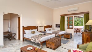 A hotel room with two beds, a sitting area, a wooden table, and a view of greenery outside. - Hotel Dev Vilas (Sawai Madhopur)