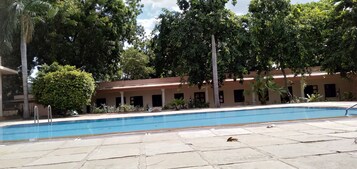 Una piscina con bordeado de baldosas, rodeada de árboles y un edificio con techo rojo.