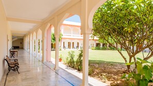 A covered corridor with marble flooring, benches, and a view of a building with arches and a lawn. - Hotel Dev Vilas (Sawai Madhopur)