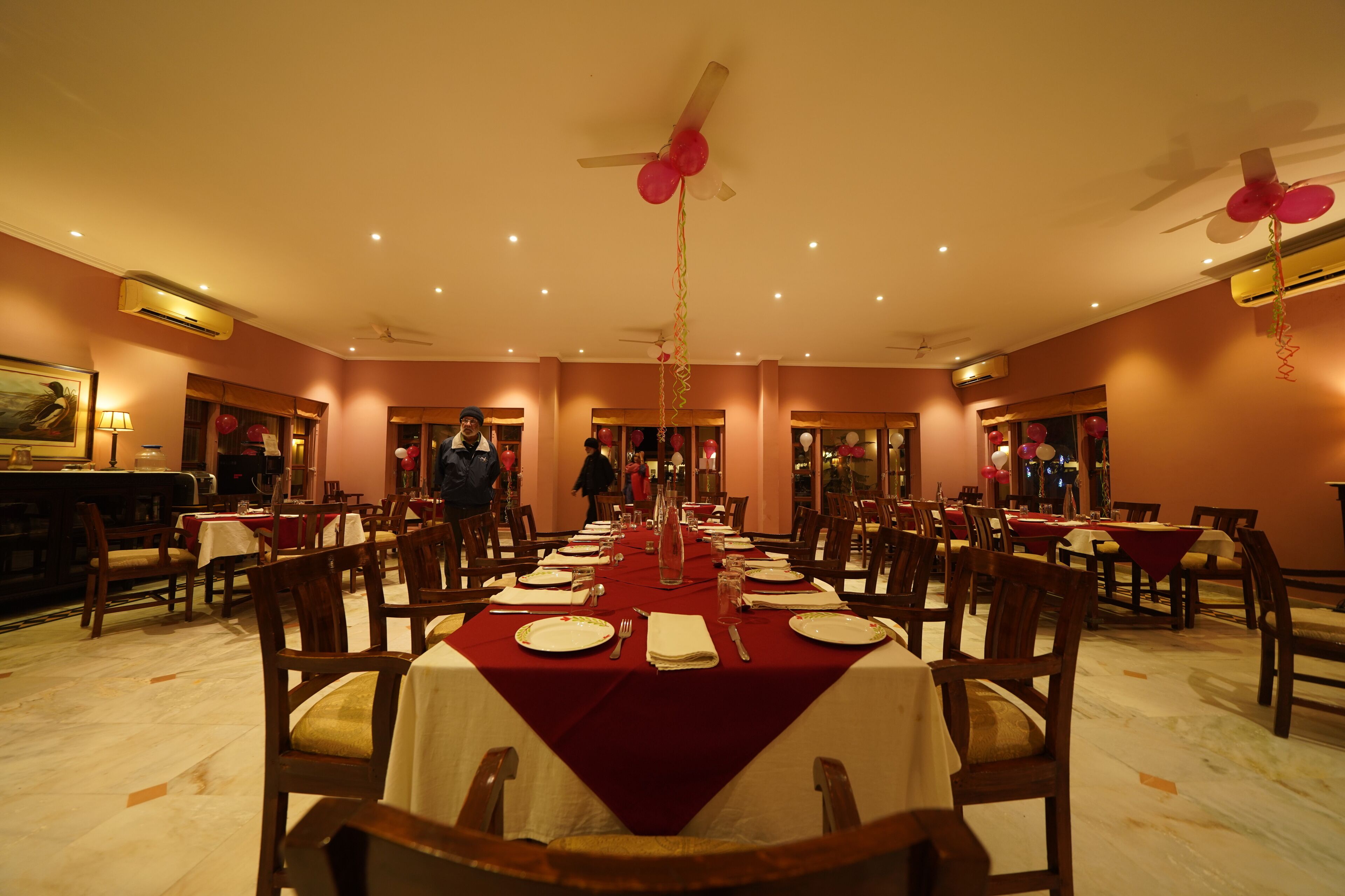 Uma sala de jantar com mesas postas para refeição, cadeiras, ventiladores de teto e balões decorativos.