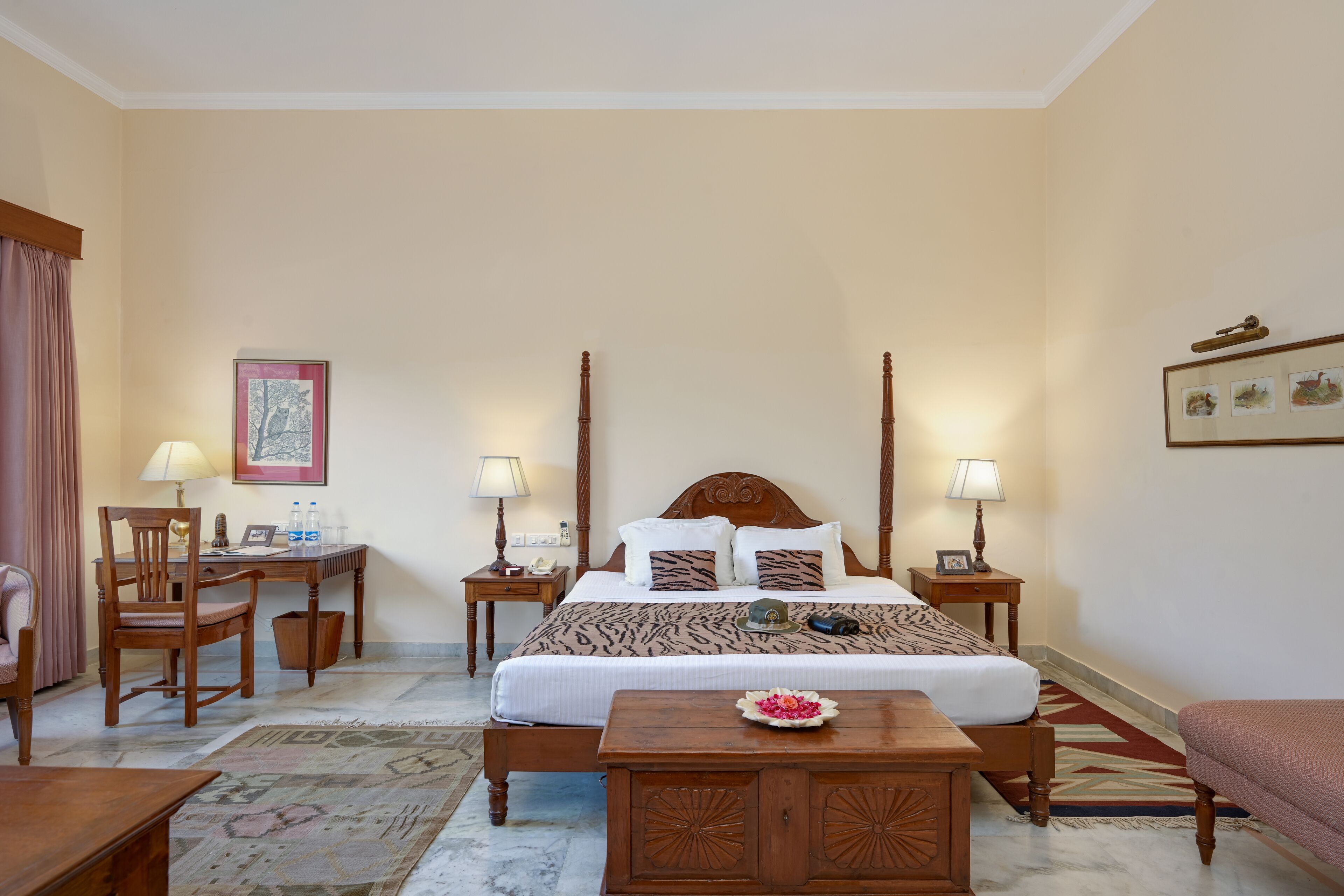 Una habitación de hotel con una cama grande, dos mesitas de noche con lámparas, un escritorio de madera, una silla y una pequeña mesa con un arreglo floral.