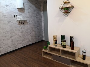 Habitación de hotel con una pared decorada con nombres de cafés parisinos, un estante de madera con botellas y una planta pequeña.