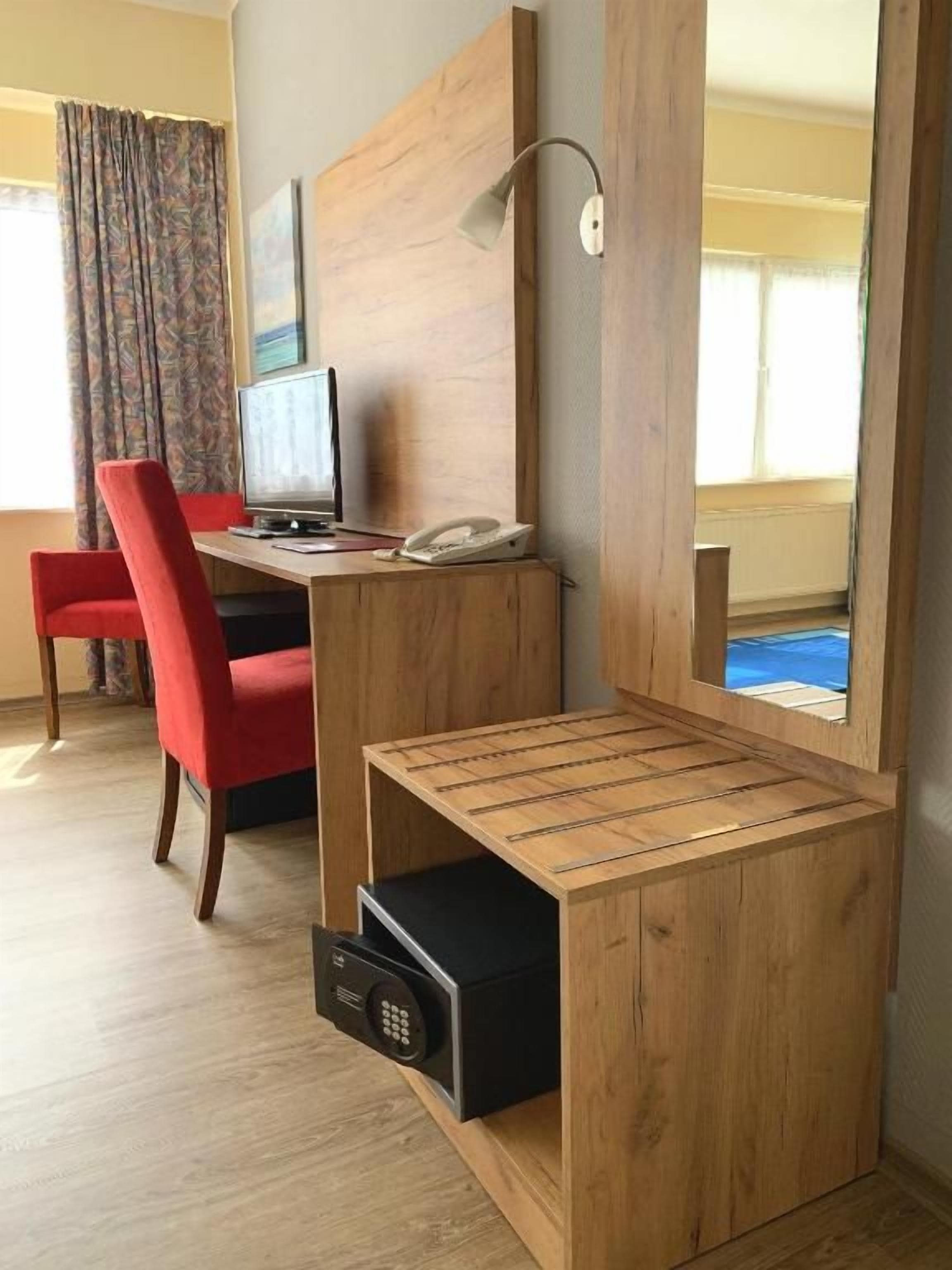 A hotel room with a wooden desk, a red chair, a television, and a mirror.