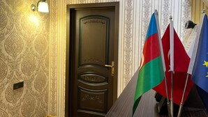 A room with a patterned wallpaper, a dark wooden door, and three flags on a stand. - VM Hostel (Ganja)