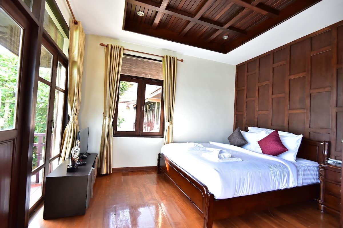 A bedroom with a wooden bed, a bedside table, a window with curtains, and a view of greenery outside.