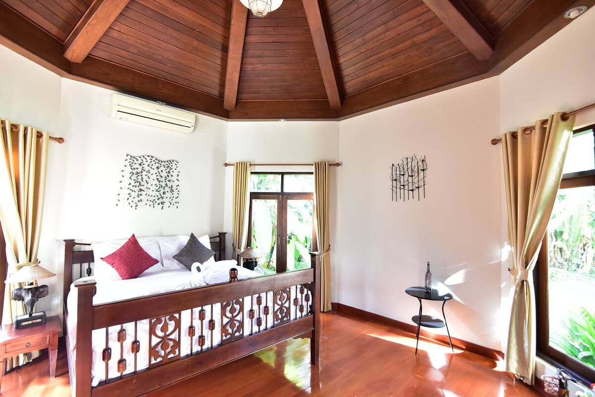 A bedroom with a wooden ceiling, a bed with a headboard, a small table, and a window with curtains.