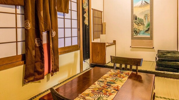 Una habitación japonesa tradicional con suelo de tatami, una mesa baja con camino de mesa floral, una silla de madera y una puerta corredera con diseños decorativos.