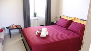 Una cama bien hecha con colcha roja, una mesita con cafetera y un florero, y una ventana con cortinas.