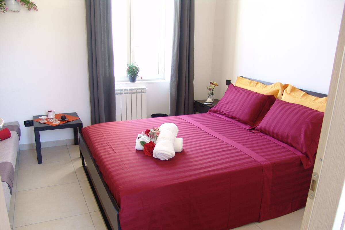 Una cama bien hecha con colcha roja, una mesita con cafetera y un florero, y una ventana con cortinas.