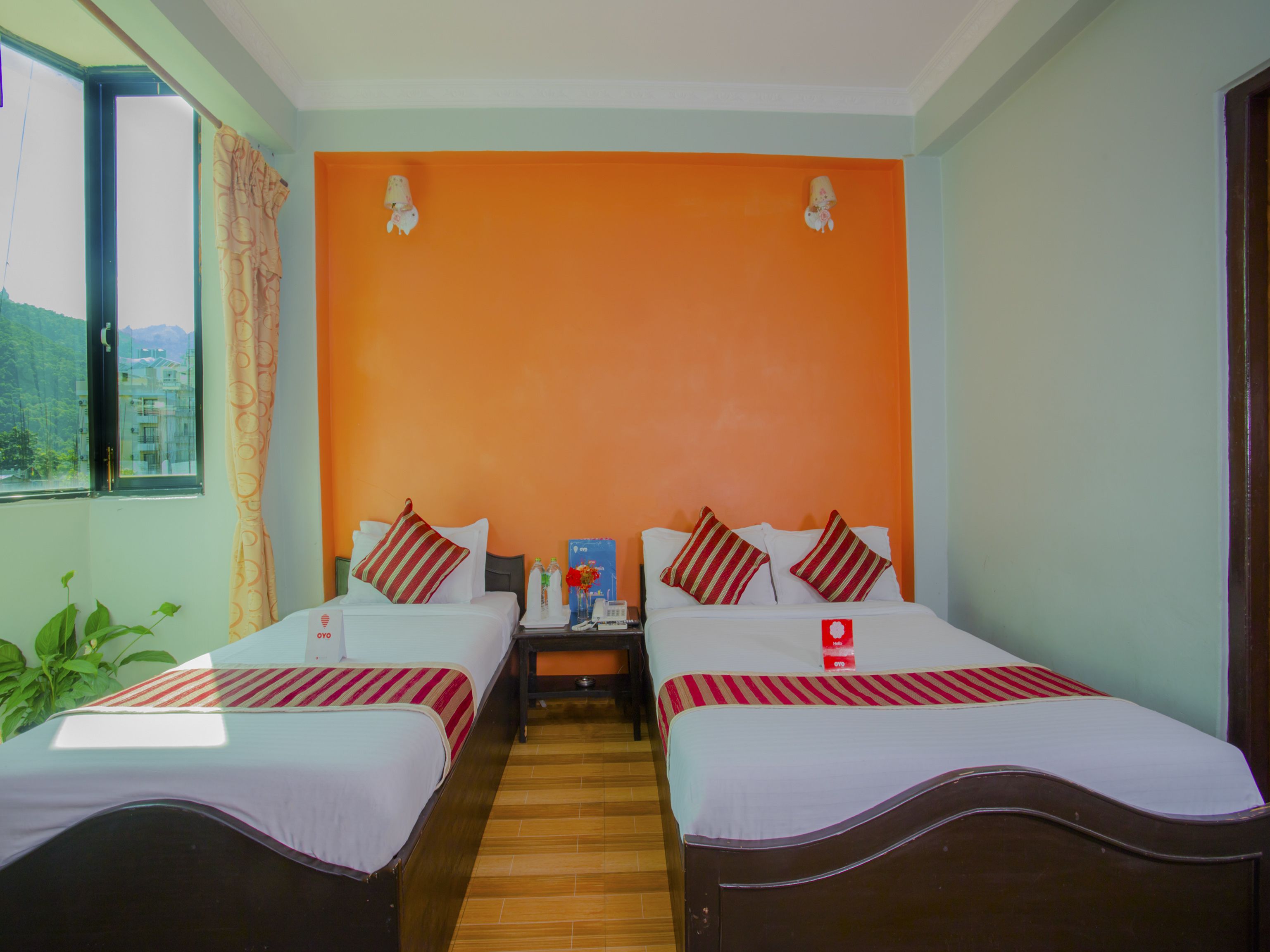 Quarto de hotel com duas camas, piso de madeira, vista para montanhas pela janela e uma parede laranja vibrante.