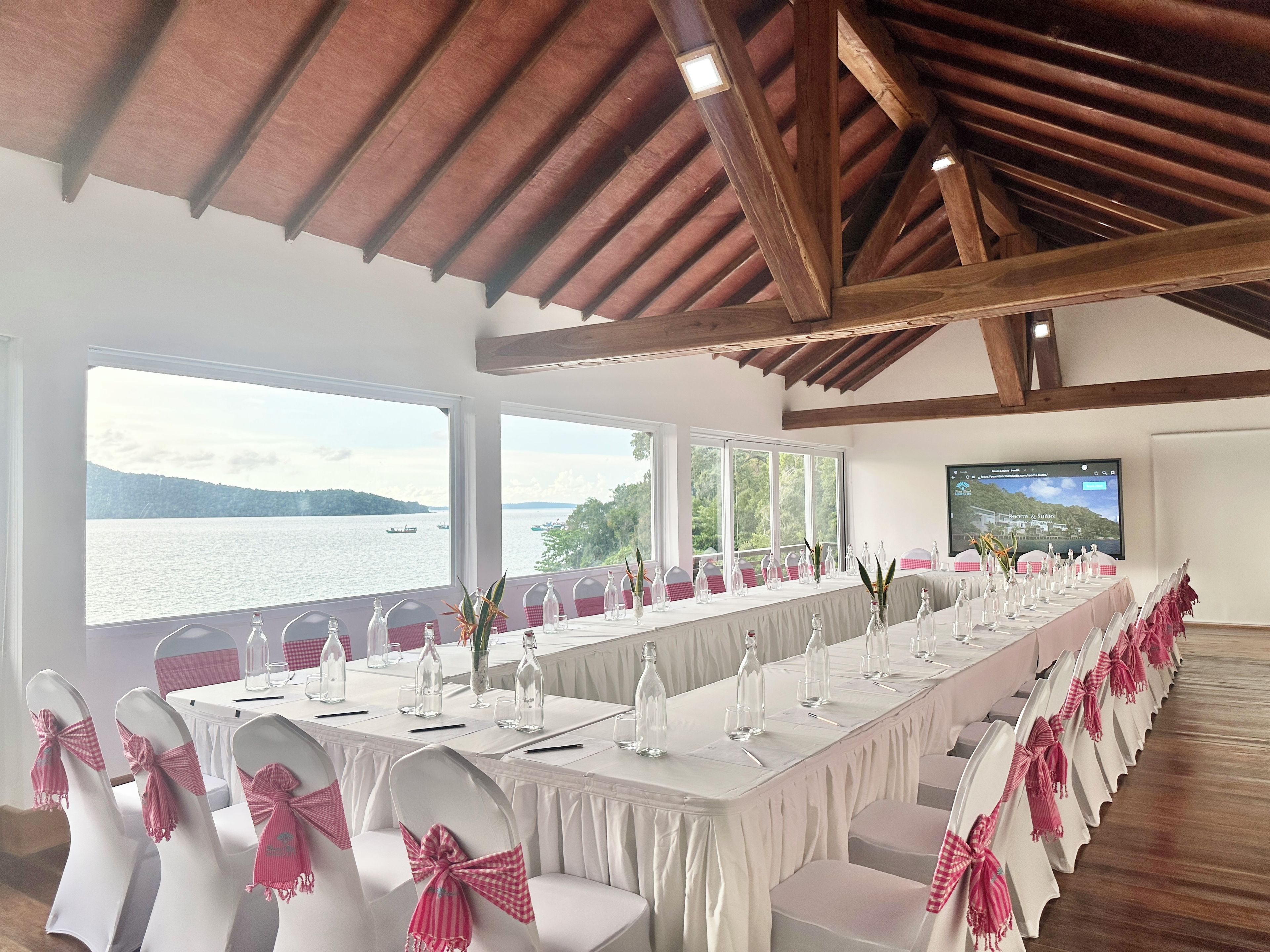 Ein Konferenzraum mit einem langen Tisch, weißen Stühlen mit rosa Schleifen und großen Fenstern mit Blick auf ein Gewässer.
