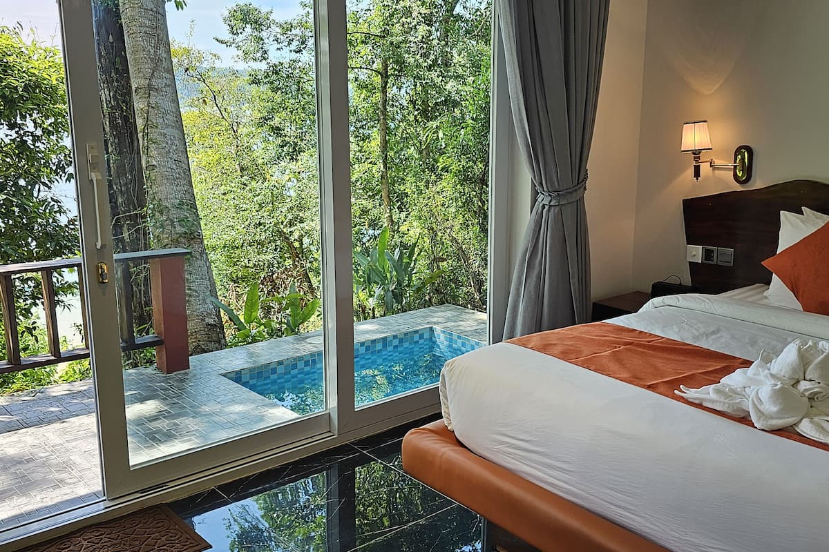 A hotel room with a bed, a nightstand, a lamp, and a view of a swimming pool and greenery.