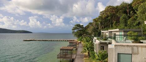 A coastal hotel with a wooden pier leading to overwater bungalows, a clear view of the sea, and lush greenery.