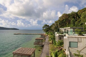 Un hotel costero con un muelle de madera que conduce a bungalows sobre el agua, vista clara del mar y abundante vegetación.