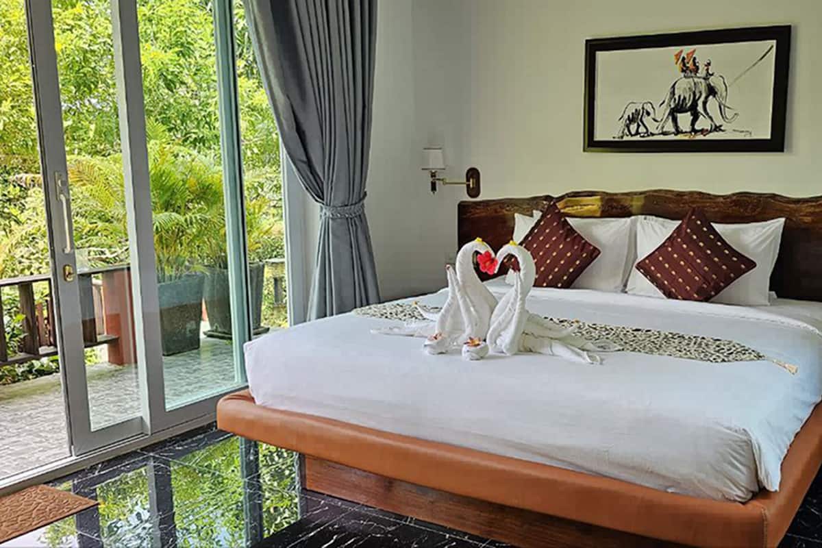 A hotel room with a large bed, a view of greenery through the glass door, and a framed picture on the wall.