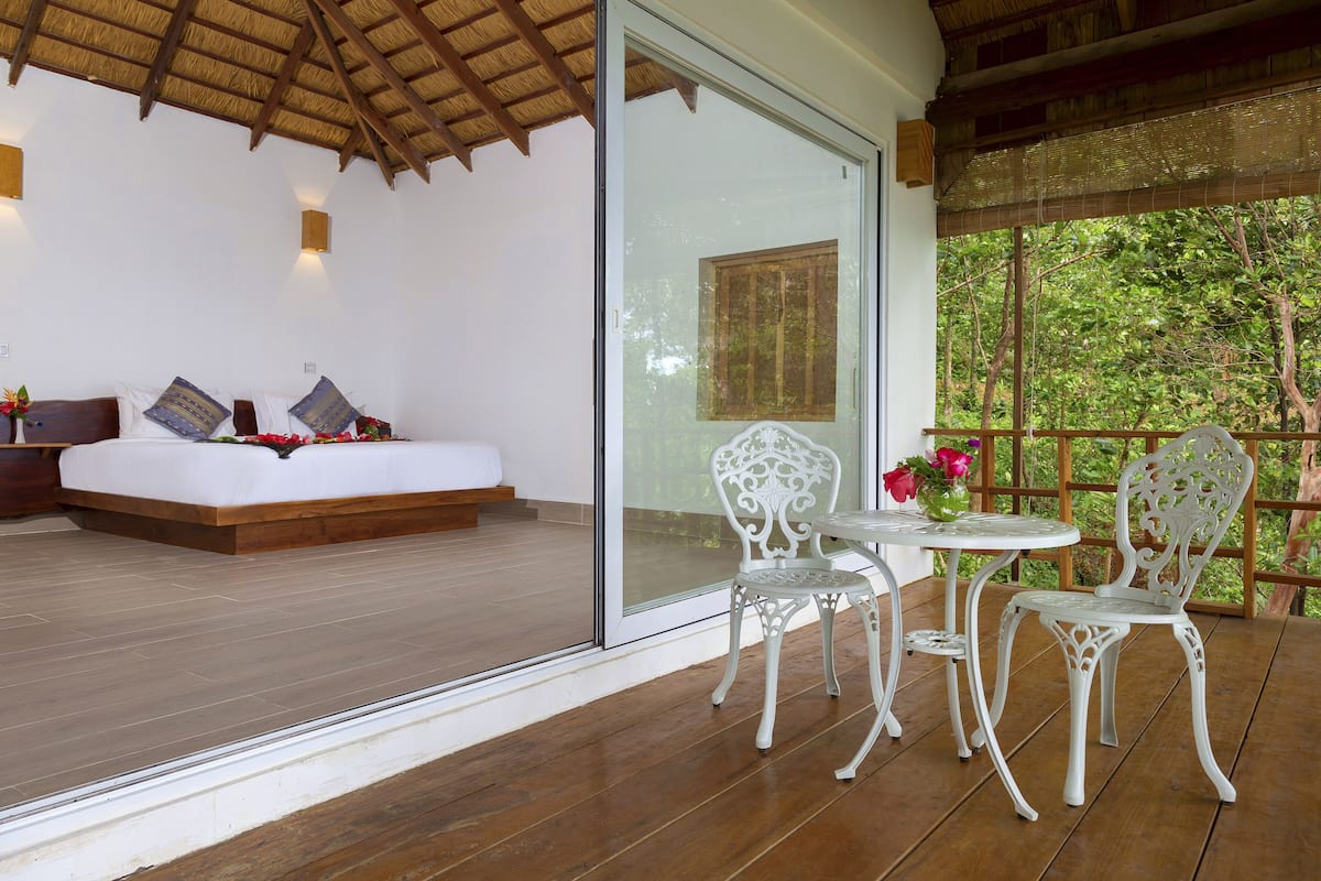 A bedroom with a large glass door leading to a balcony, a bed with pillows, and a small table with chairs.