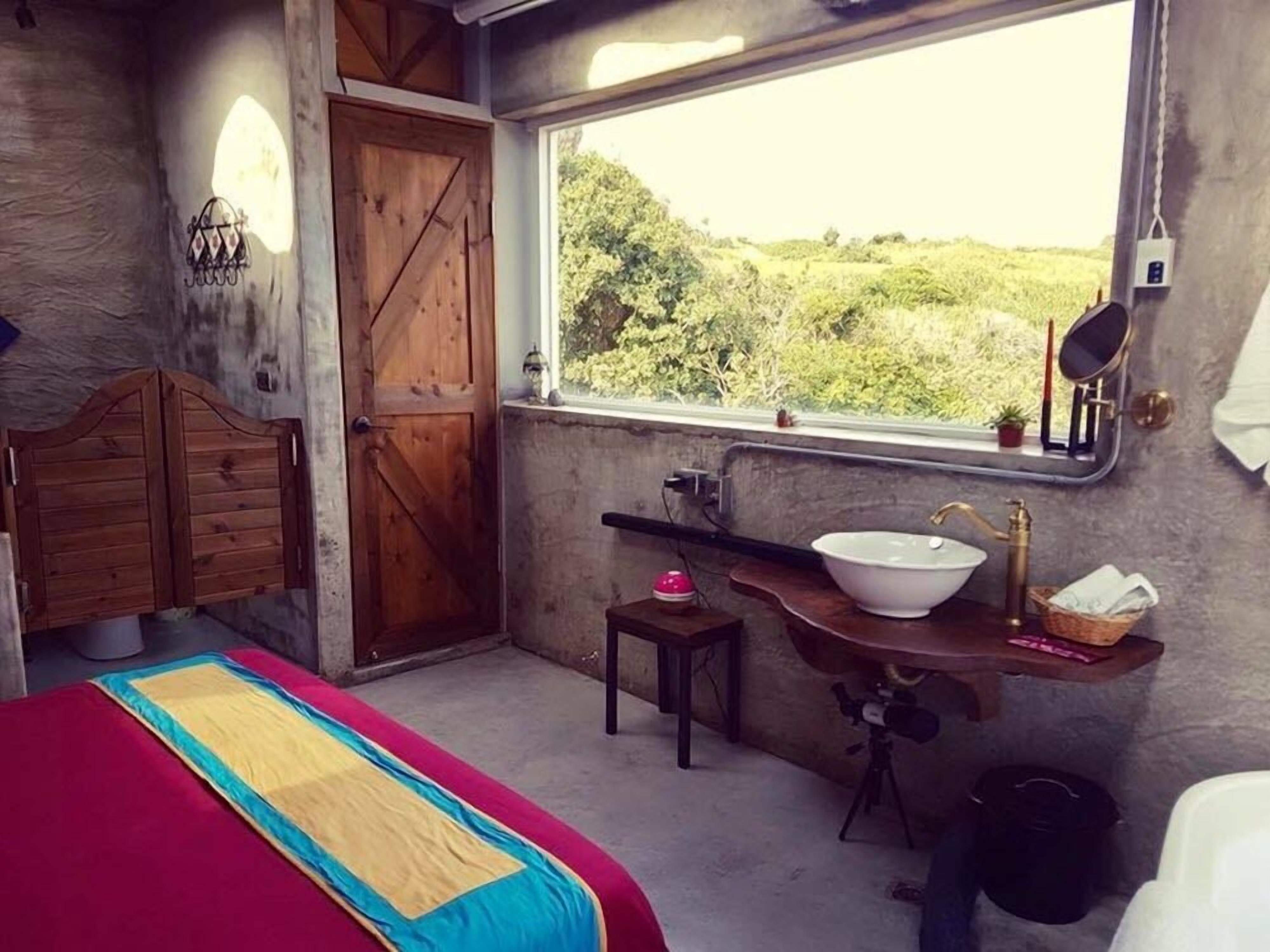 A bedroom with a large window, a wooden door, a bed with a colorful bedspread, a small bedside table with a lamp, and a bathroom with a sink and mirror.