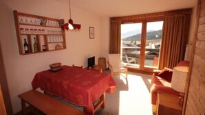 Ein Zimmer mit einem Bett, einem Tisch und einer Bank. Ein Regal mit Flaschen und Gläsern ist vorhanden. Ein Stuhl steht in der Nähe eines Fensters mit Vorhängen.