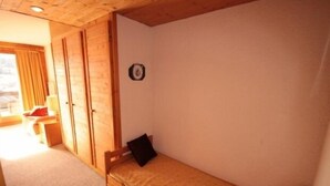 Ein kleines Zimmer mit einem Einzelbett, einem hölzernen Bettgestell, einem Nachttisch, einem Stuhl und einem Fenster mit Vorhängen.