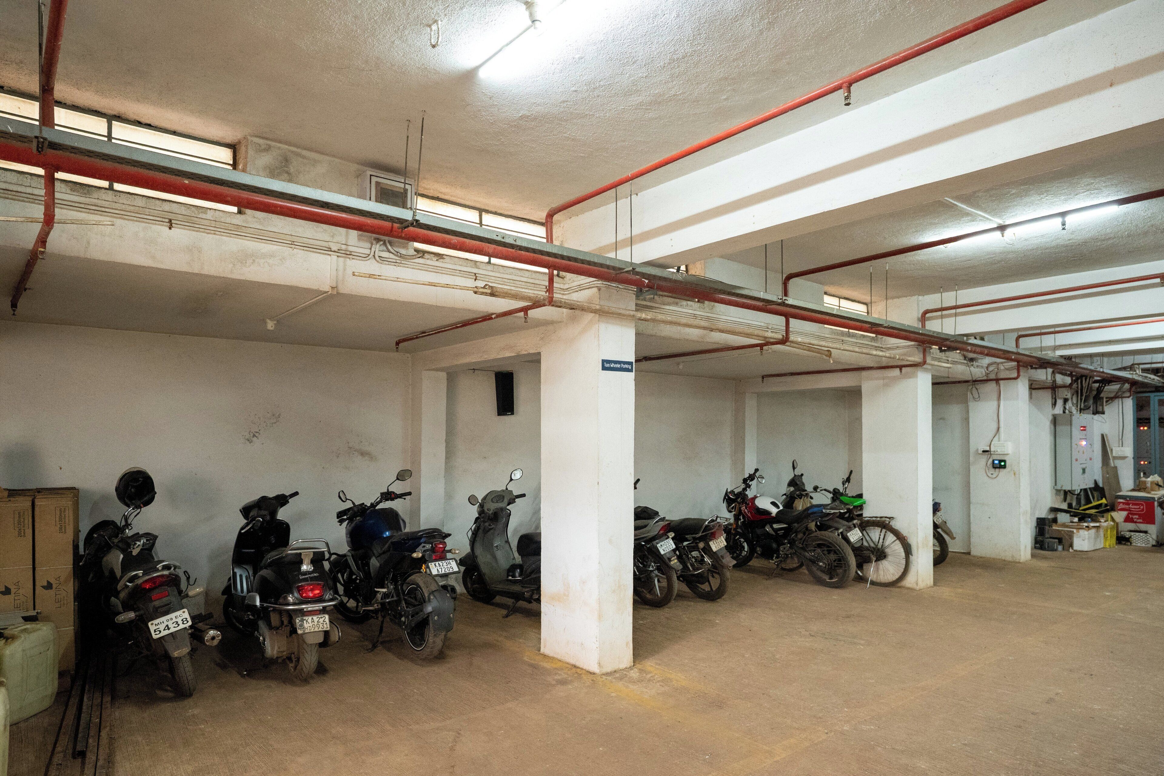 A parking garage with multiple motorcycles parked in rows, concrete pillars, and overhead pipes.