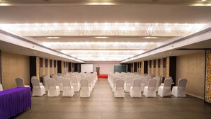 Un salón de conferencias con filas de sillas blancas, un escenario con un atril y una pantalla grande.