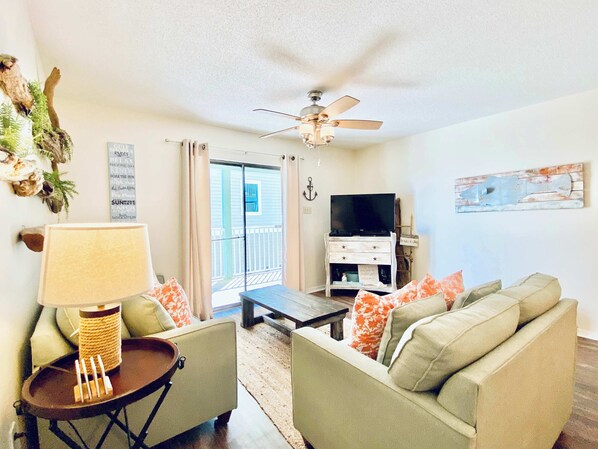 A living room with a sofa, armchair, and a small table with a lamp. There is a television on a stand and a ceiling fan. - Sundial 2C by ALBVR - Beachfront Complex - Great Location and Amenities (Gulf Shores)