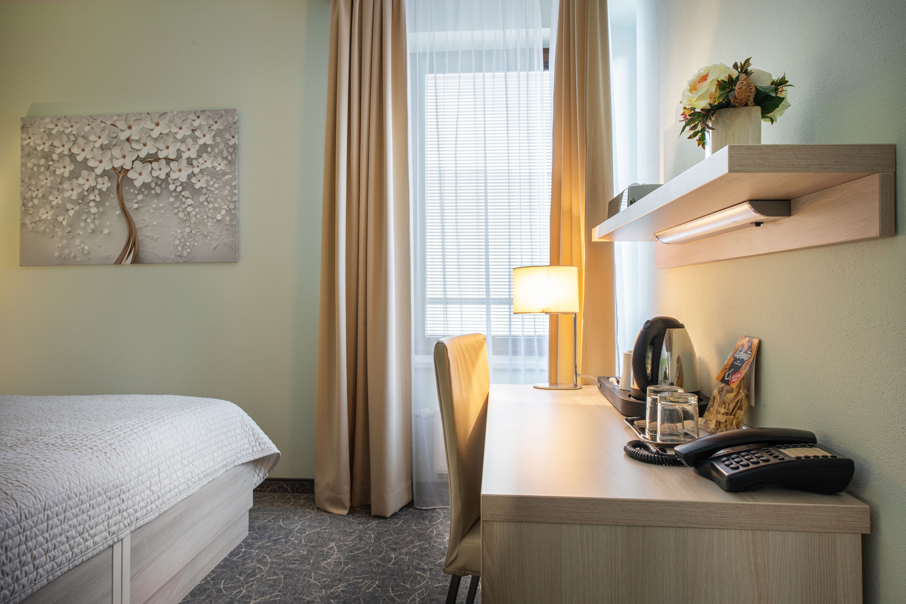 Una habitación de hotel con una cama, un escritorio con lámpara, un teléfono y una estantería de pared con un jarrón de flores.