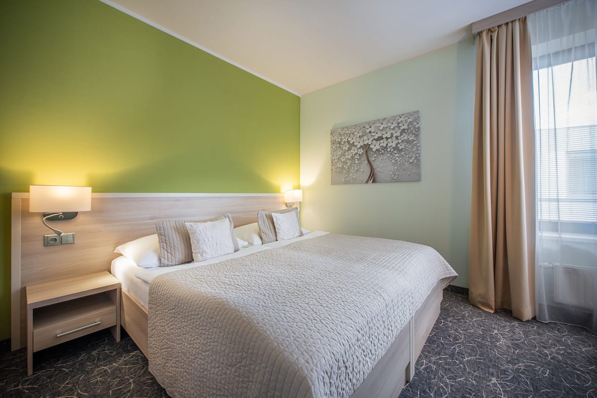 Una habitación de hotel con una cama grande, una mesita de noche, una lámpara de pared y un ventanal con cortinas.