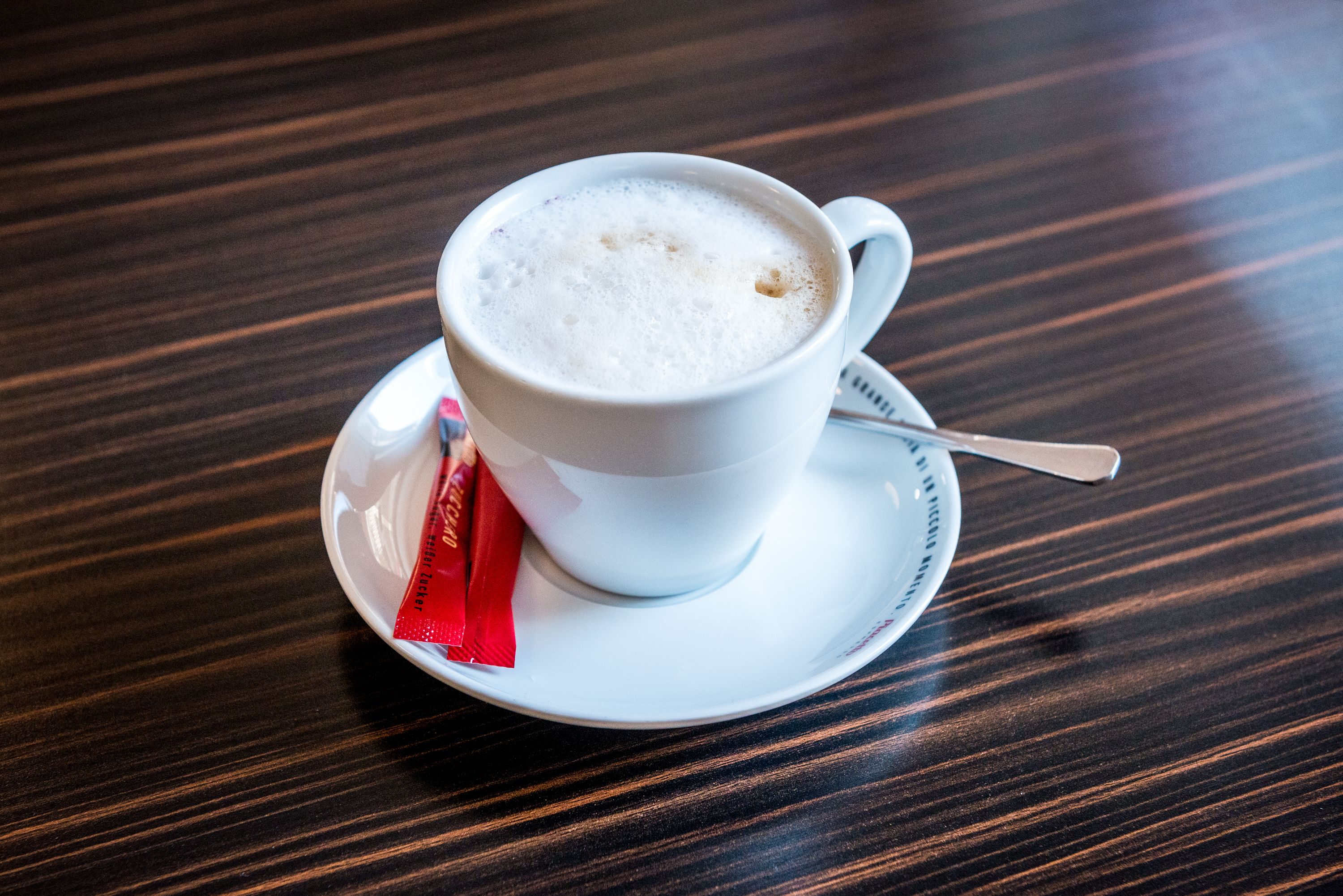 Una taza blanca de café con espuma, sobre un platito blanco con una cuchara, sobre una mesa de madera.