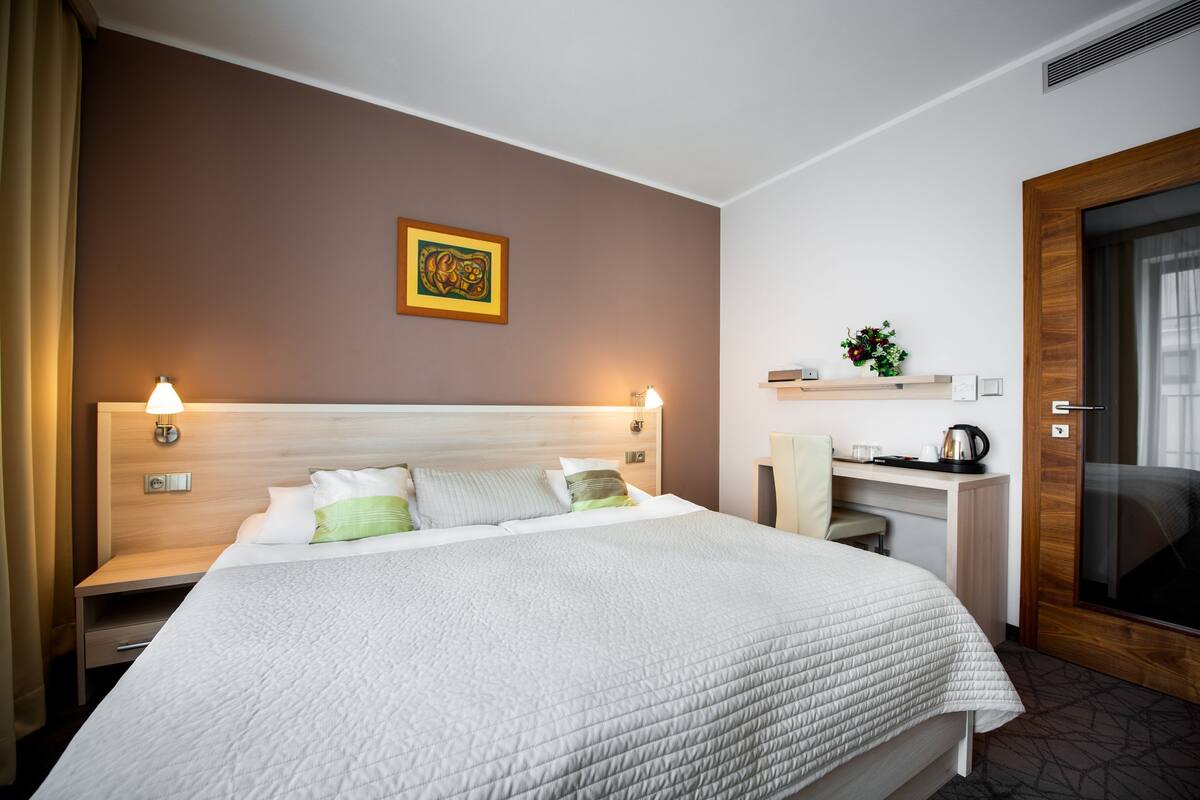 Habitación de hotel con una cama grande, lámparas de noche, un escritorio con silla, una planta pequeña y una puerta.