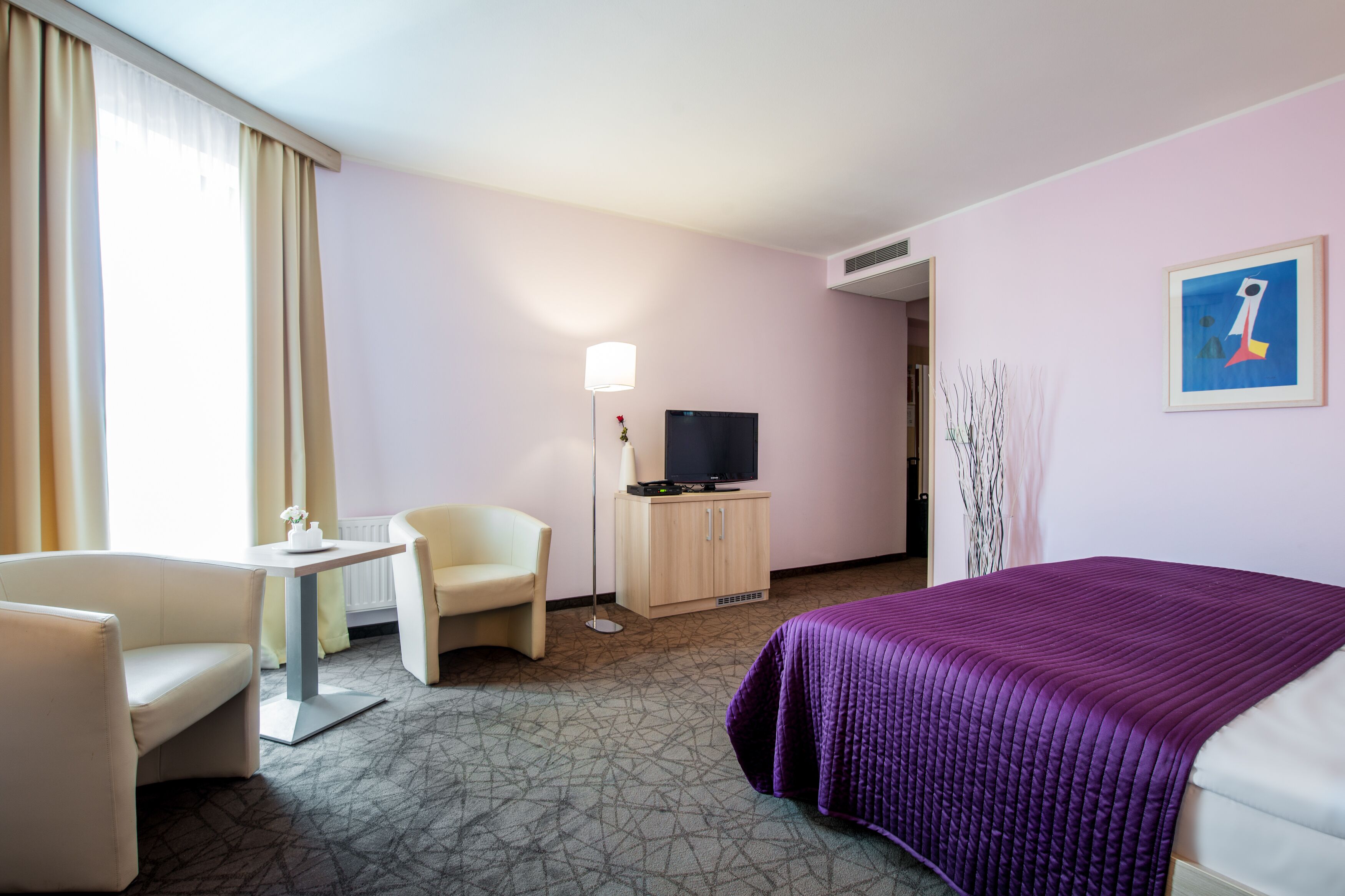 A hotel room with a bed, a desk, a chair, a television, and a painting on the wall.