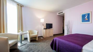 A hotel room with a bed, a desk, a chair, a television, and a painting on the wall.