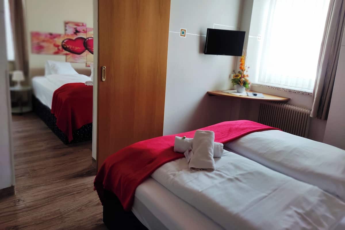 Une chambre d’hôtel avec deux lits, un téléviseur fixé au mur, une fenêtre avec des rideaux et une couverture rouge sur l’un des lits.