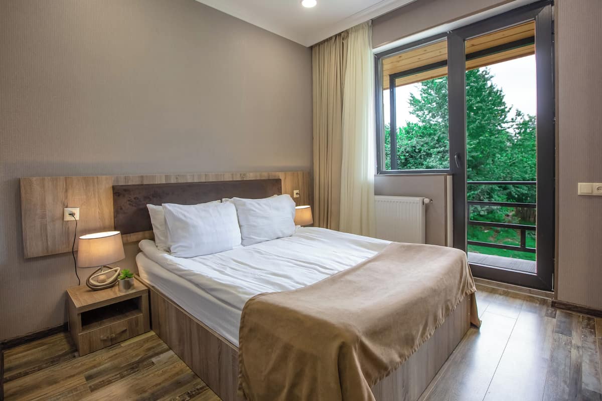 A bedroom with a bed, bedside table, lamp, and a view of greenery through a window.