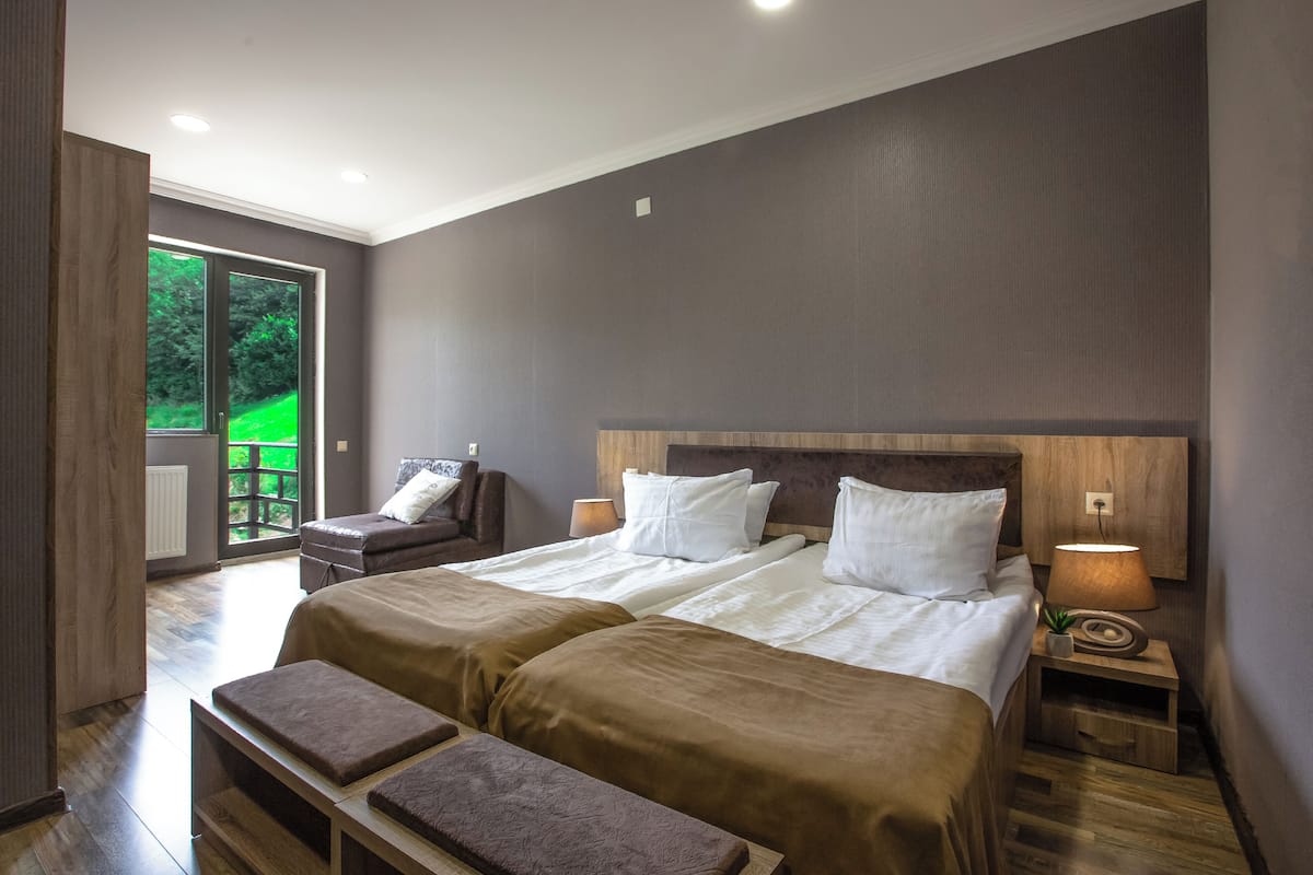 A bedroom with a large bed, two bedside tables, a bench, and a view of a green outdoor area.