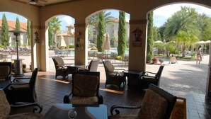 An outdoor seating area with wicker chairs and tables, a pool in the background, and palm trees. - Gorgeous Condo in Prestigious Championsgate Golf Resort Community w View of Pool (Championsgate)