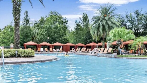 A swimming pool with lounge chairs and umbrellas, surrounded by palm trees and greenery. - Gorgeous Condo in Prestigious Championsgate Golf Resort Community w View of Pool (Championsgate)