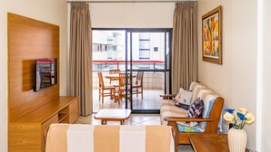 Sala de estar com sofá, televisão presa a um painel de madeira, mesa de jantar com cadeiras e varanda com vista para edifícios.