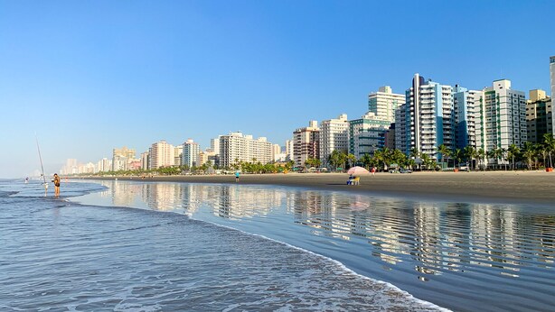 Uma praia com mar calmo, areal arenoso e o horizonte urbano ao fundo.