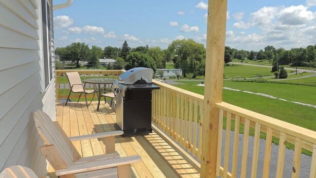 Una terraza de madera con una parrilla, sillas y vistas a un campo verde.