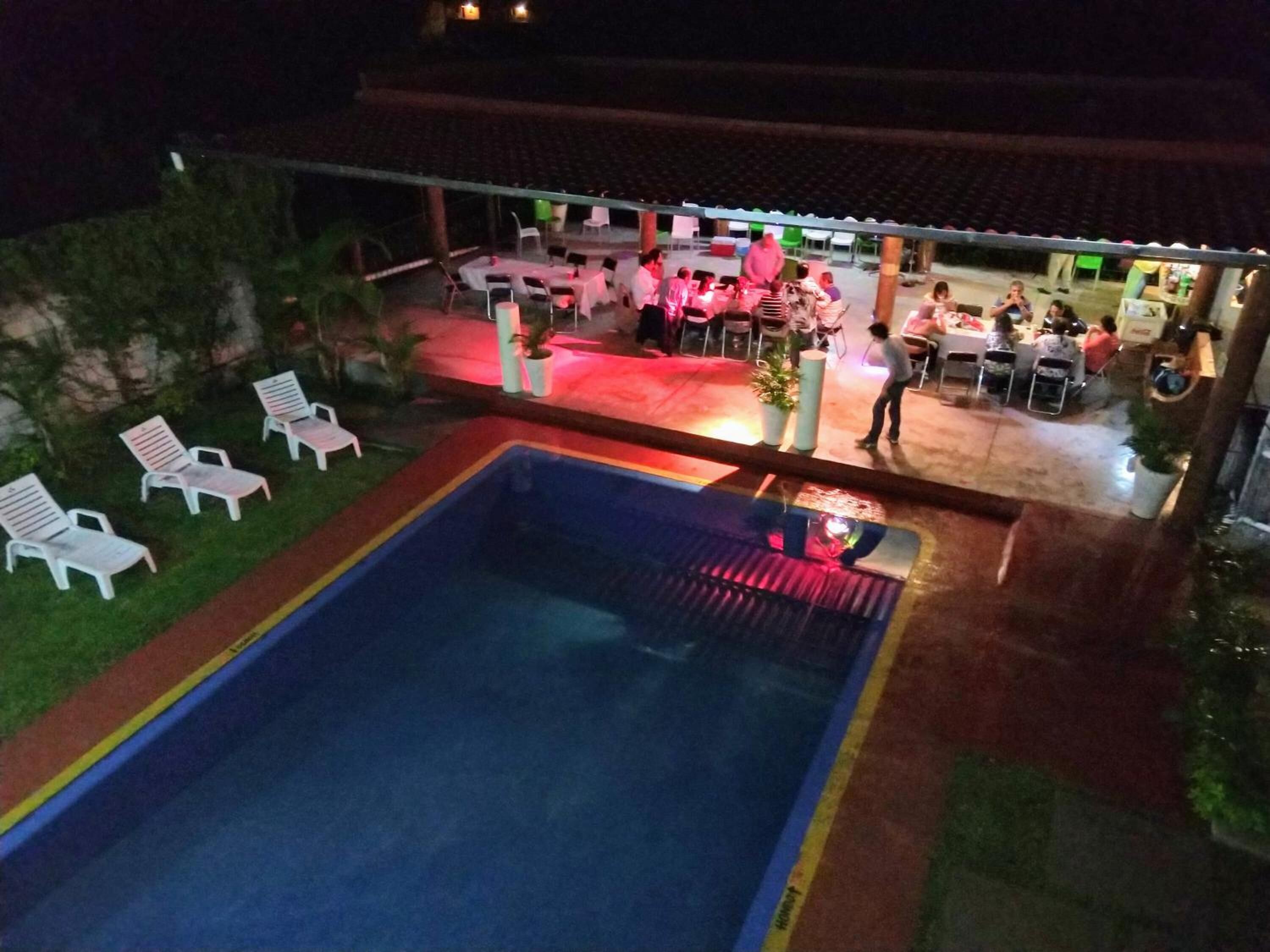 Un área de piscina al aire libre con sillones reclinables y mesas, rodeada por una zona cubierta con sillas y mesas.