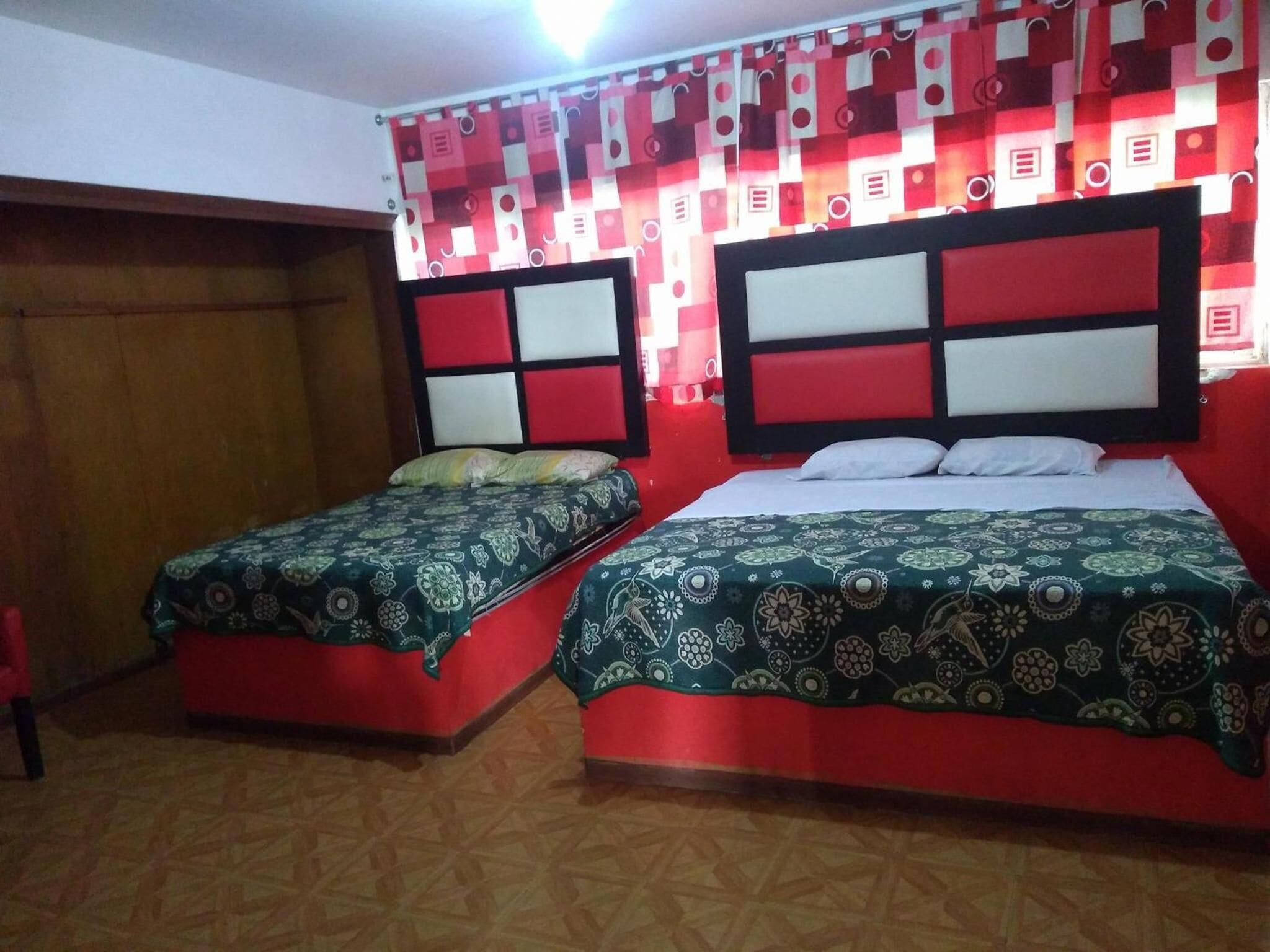 A room with two beds, each with patterned bedding, red and white headboards, and a red curtain with geometric patterns.
