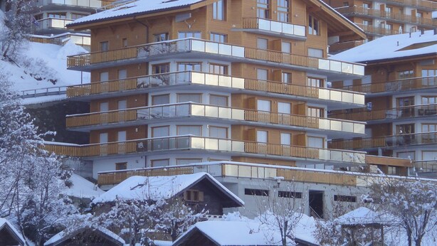 Ein mehrgeschossiges Gebäude mit Balkonen, Holzfassade und Schnee auf dem Dach und den umliegenden Bäumen.