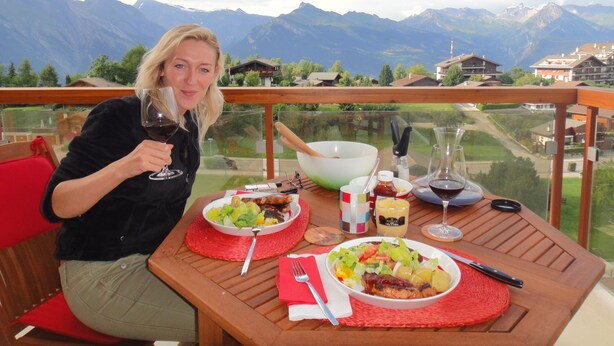 Une personne est assise à une table, devant une vue sur les montagnes, et a devant elle un saladier rempli de salade et un verre de vin rouge.